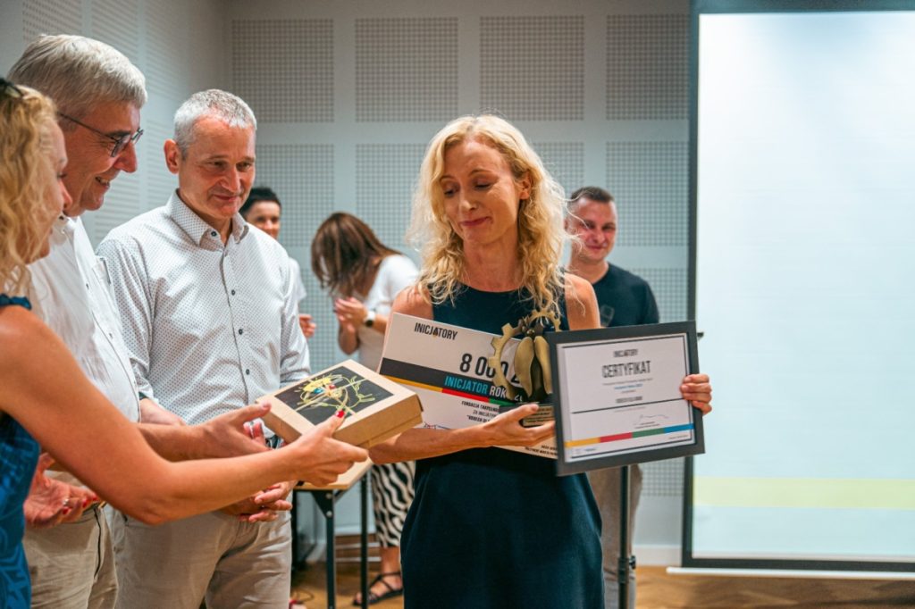 Na zdjęciu Agata Robińska odbierająca nagrodę, którą wręcza kobieta w kręconych blond włosach i mężczyźni w siwych włosach i białych koszulach, jeden z nich nosi okulary.