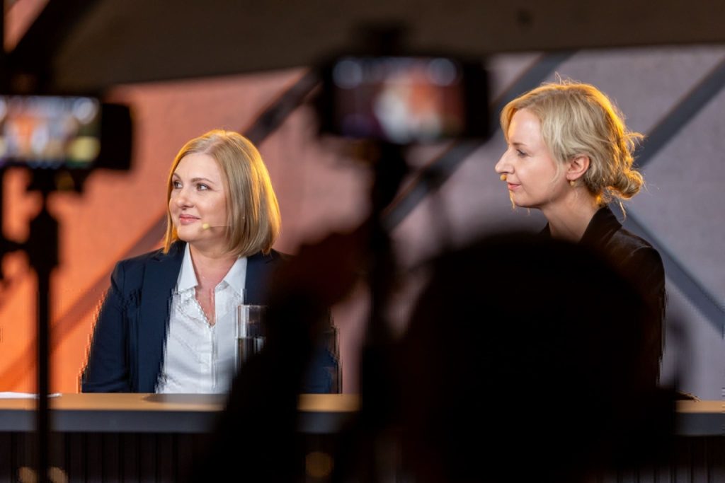 Dwie kobiety – jedna blondynka, druga szatynka – siedzą przy stole w studiu nagraniowym. W tle profesjonalne oświetlenie i sprzęt. Ujęcie zza sylwetki kamerzysty.