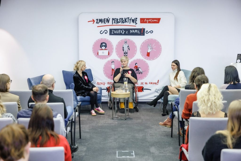 Three women on stage are having a conversation; one of them is in a wheelchair. Behind them is a banner with the slogan “Change perspective with the Agates.” The audience listens attentively.