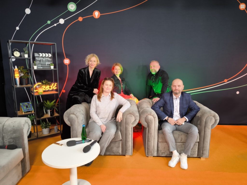 Five people pose for a group photo in a studio with a colorful backdrop and a sign reading “Cześć” (Polish for “Hi”). In the background, set decorations include books, icons, and a shelf. Microphones lie on the table.