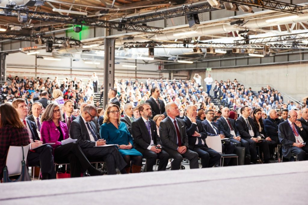 View of the auditorium during the Global Disability Summit. People in formal attire sit in the front rows. A large crowd is visible in the background, filling the stands.