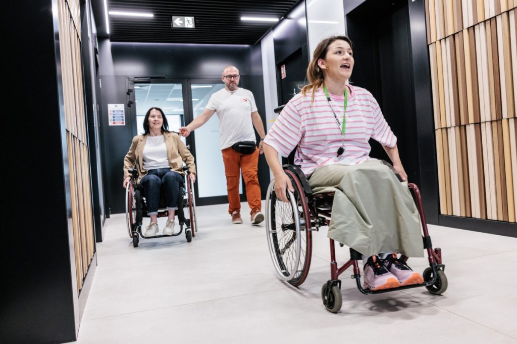 Dwie uczestniczki akcji edukacyjnej „Zmień perspektywę” poruszają się na wózkach w korytarzu biurowym centrali Leroy Merlin Polska. Za nimi idzie mężczyzna wspierający ćwiczenie. Symulacja pozwala doświadczyć mobilności i barier architektonicznych.