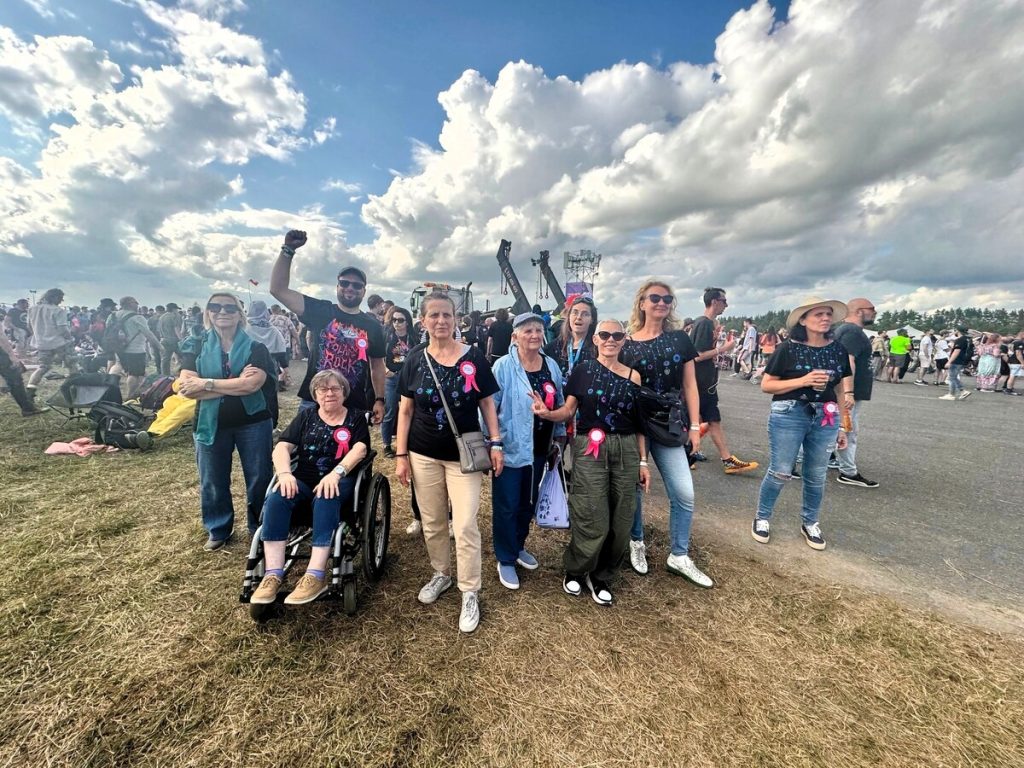 Nine women and one man pose in front of the main festival area. A large crowd and a stage are visible in the background. Some people wear T-shirts with the “Pol’and’Rock” logo.