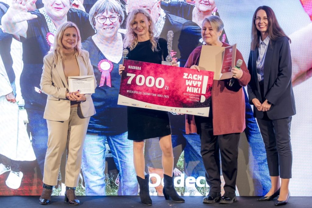 Agata Robińska i Anna Nowak stoją na scenie z dwiema kobietami podczas gali. Trzymają statuetkę i symboliczny czek na 7000 zł z napisem „Zachwytnik Roku 2025”. W tle ekran z grupowym zdjęciem uczestniczek projektu “Oddech dla Mam”.