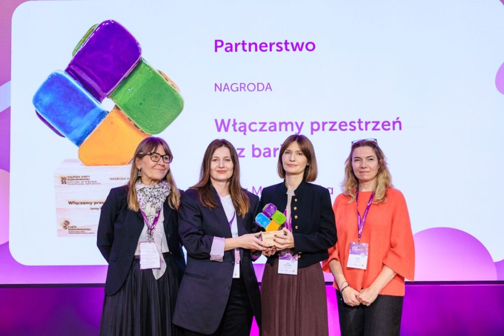 Four women standing together on stage during the Diversity Charter gala. Two of them are holding a colorful award statuette. In the background, a screen reads: “Partnership. Award. We create barrier-free spaces. Leroy Merlin Polska.”