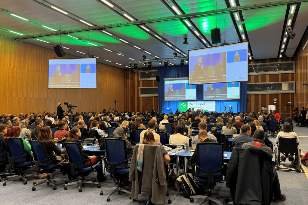 The audience sits at tables during the Zero Project conference at the UN headquarters in Vienna. On stage, there is a panel discussion, above which are two large screens broadcasting the speeches. The Zero Project logo and stage lighting are visible.