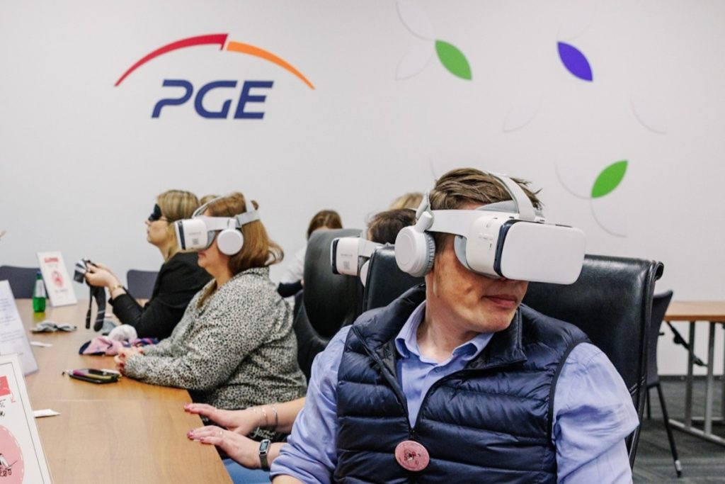 Four people sit at a conference table wearing VR headsets, experiencing neurodiversity. In the background there is a wall with the PGE logo.