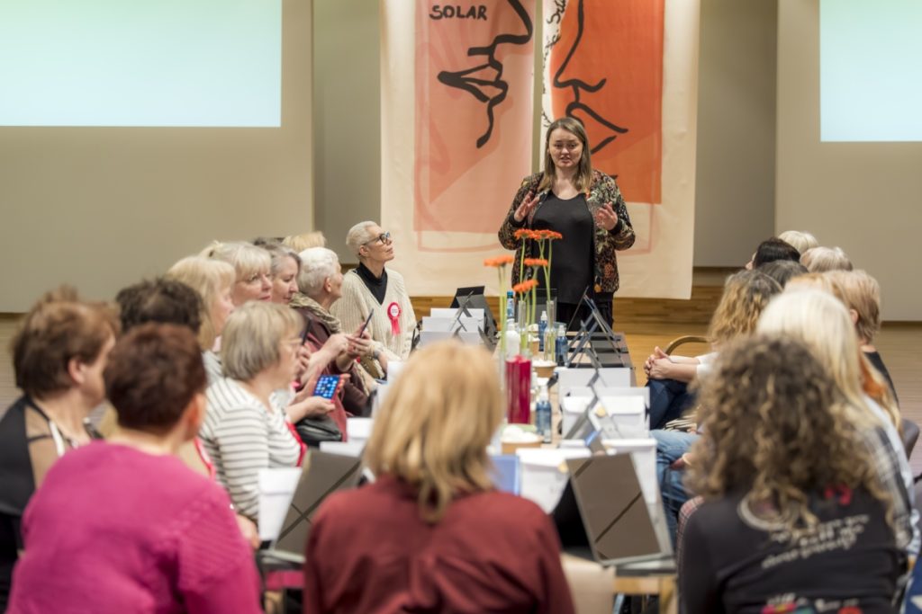 Women gathered around a long table during a “Breath for Moms” workshop, with one woman standing at the front and speaking to the group in a warm, supportive setting.