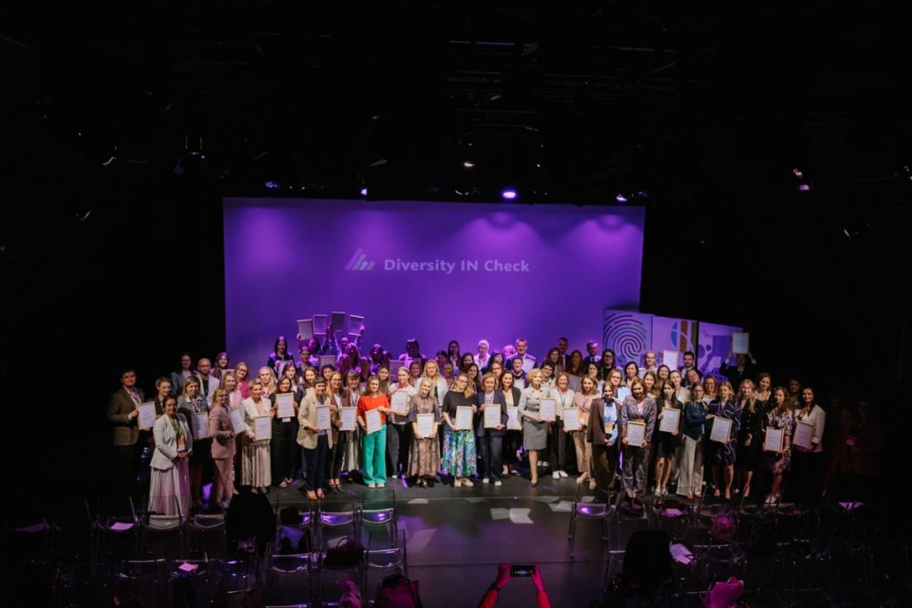 Grupa osób stoi na scenie podczas wydarzenia Diversity IN Check. Większość trzyma oprawione certyfikaty, a sala jest oświetlona fioletowym światłem.