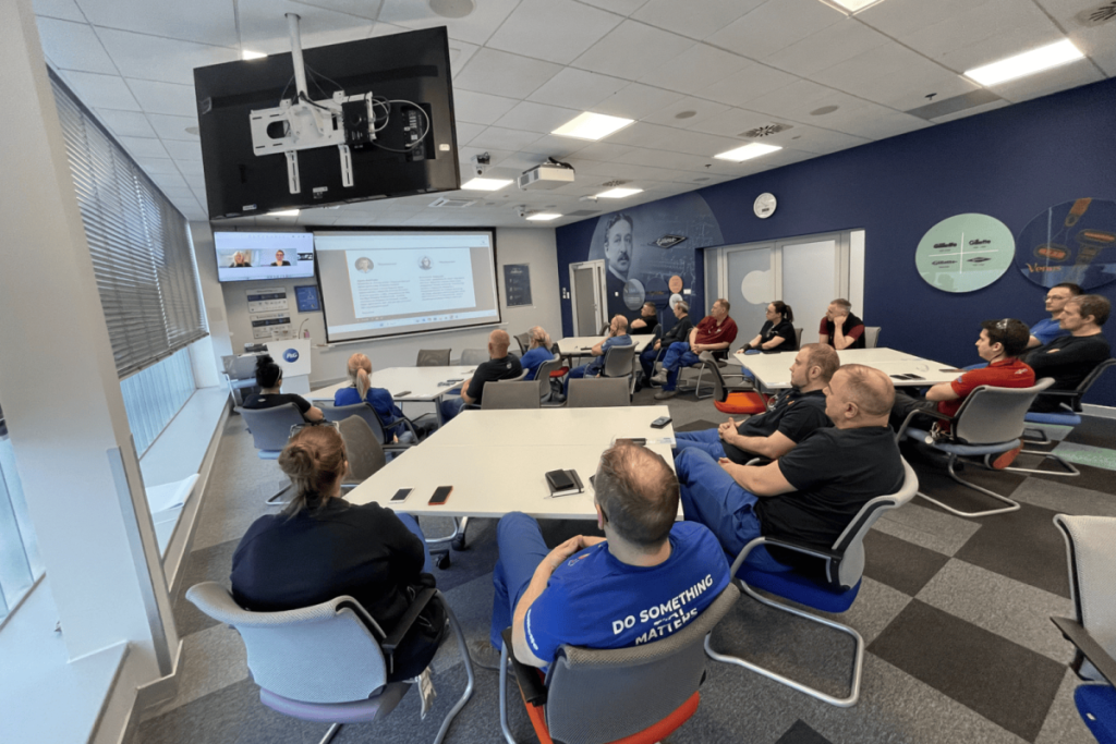 Participants in the hybrid workshops sit at tables in a P&G conference room and watch a presentation displayed on a screen.